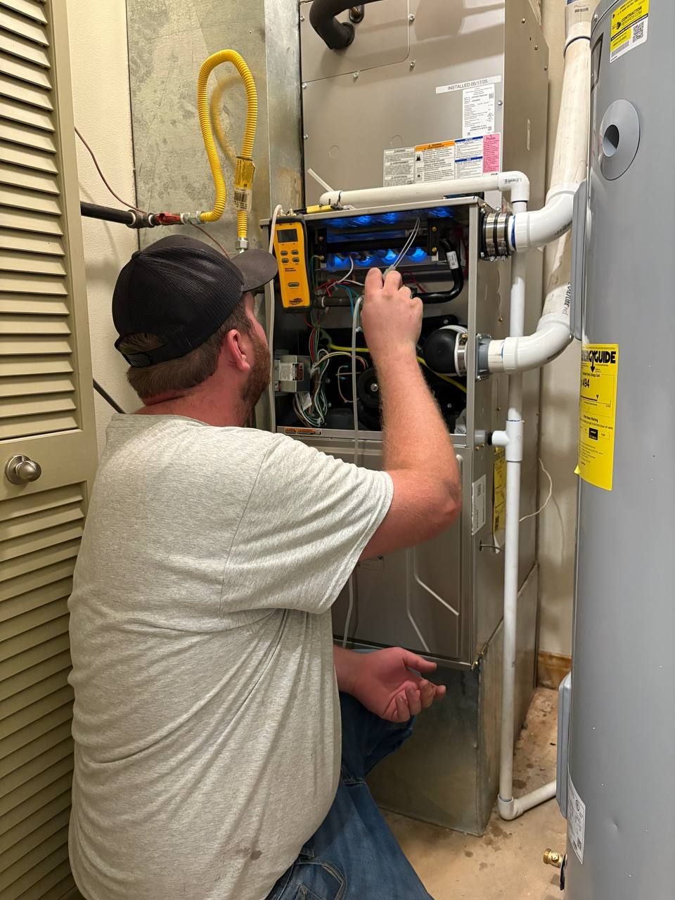 Technician repairing a furnace with various tools and wires visible, wearing a cap and gray shirt.